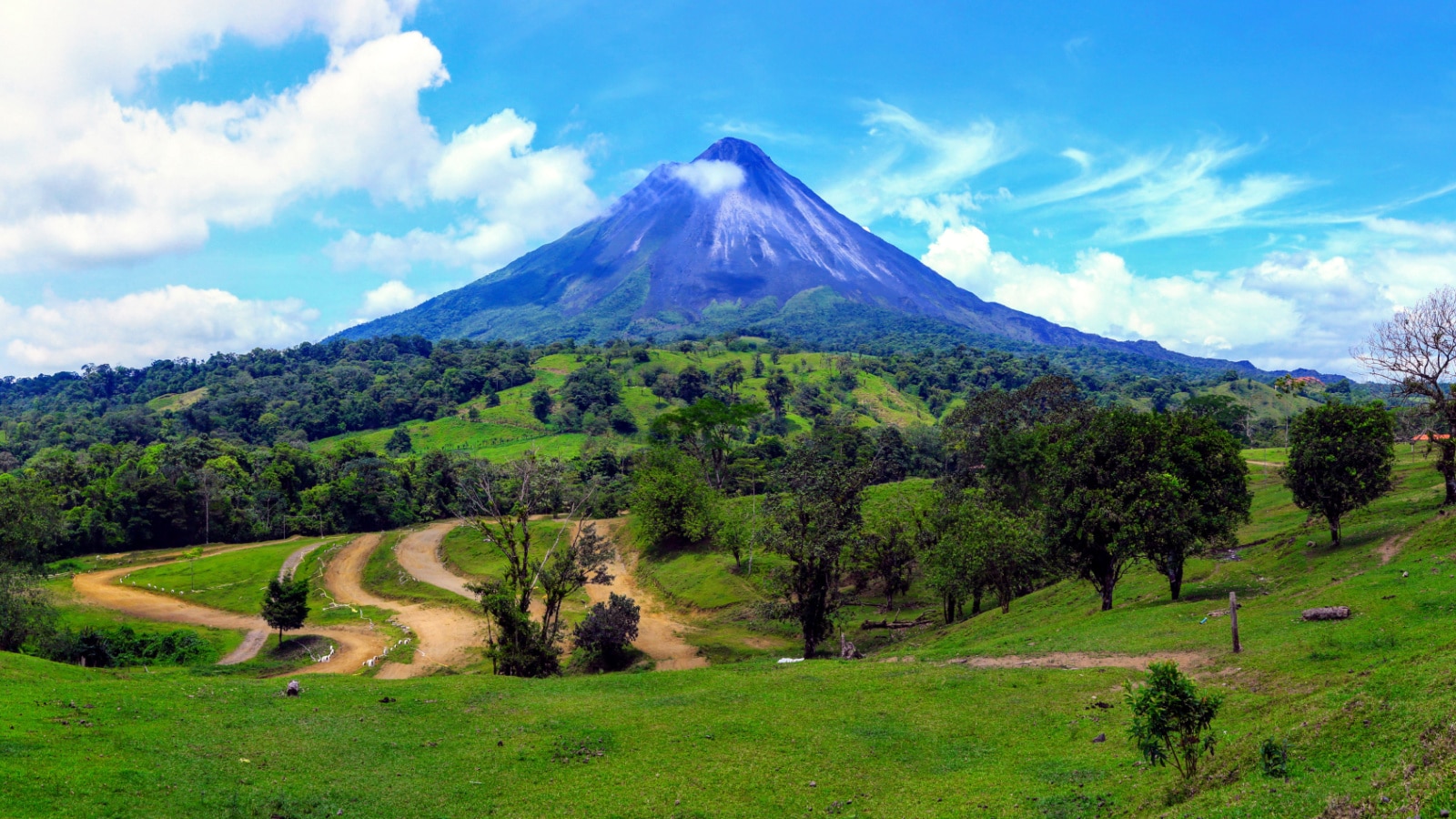 A landscape photo of Arenal volcano in Costa Rica on a clear day. The large mountain towers in the background, behind green fields filled with trees and hills.