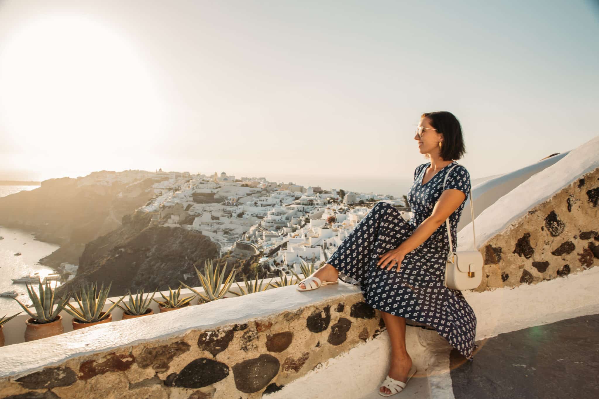 Lindsey wearing a navy Karina Midi dress with short sleeves, sitting on a ledge overlooking the white homes of Oia, Santorini, Greece