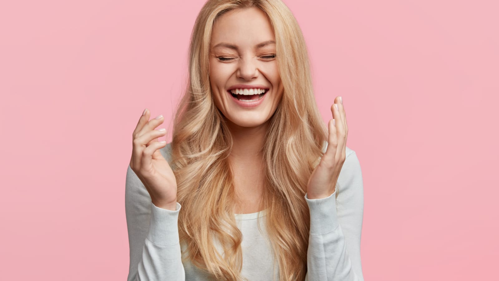 Isolated shot of joyful blonde young cute woman laughs joyfully as hears funny anecdote from friend, has long light hair, poses against pink studio wall. Happiness and positive emotions concept