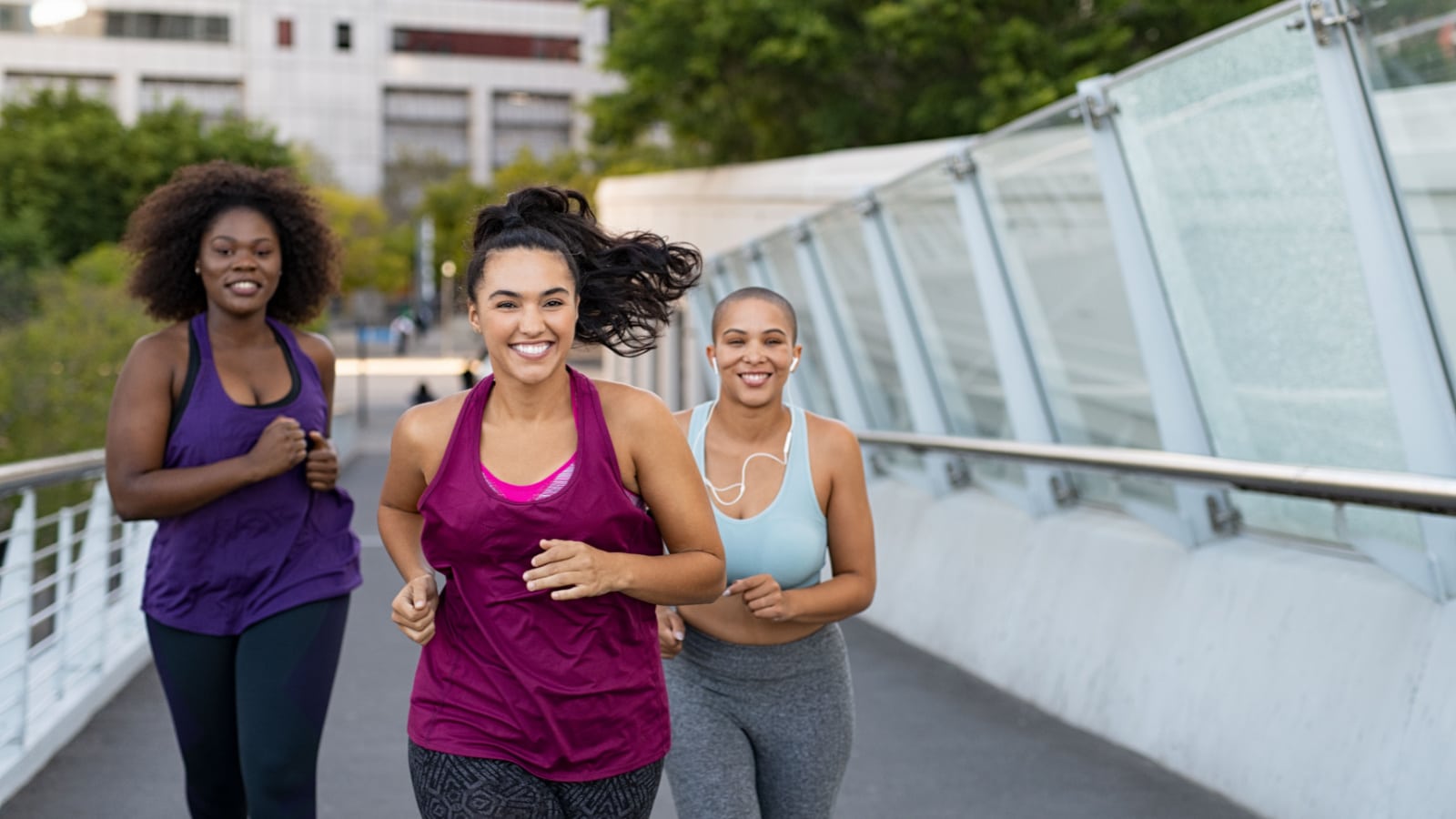 Happy young curvy women jogging together on city bridge. Healthy girls friends running on the city street to lose weight. Group of multiethnic oversize women running with building in the background.