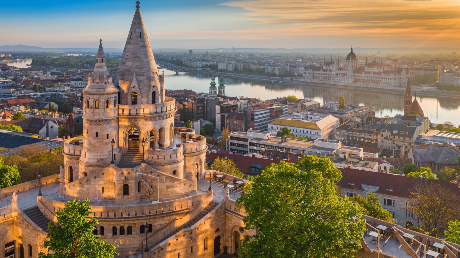 An aerial photo of Budapest, Hungary showing a beautiful golden summer sunrise with the tower of Fisherman's Bastion and green trees in the foreground. Parliament of Hungary and River Danube is in the background under blue skies.