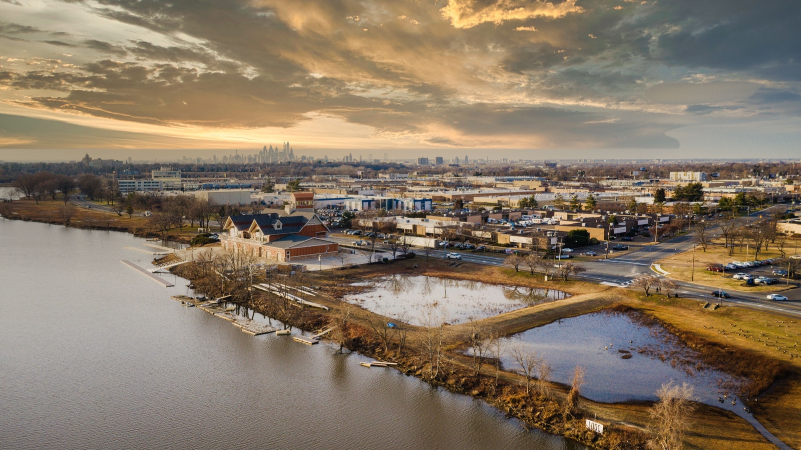 An aerial photo of Camden, New Jersey at sunset, showing a quiet town with parks on the riverfront and the New York city skyline in the distant background.