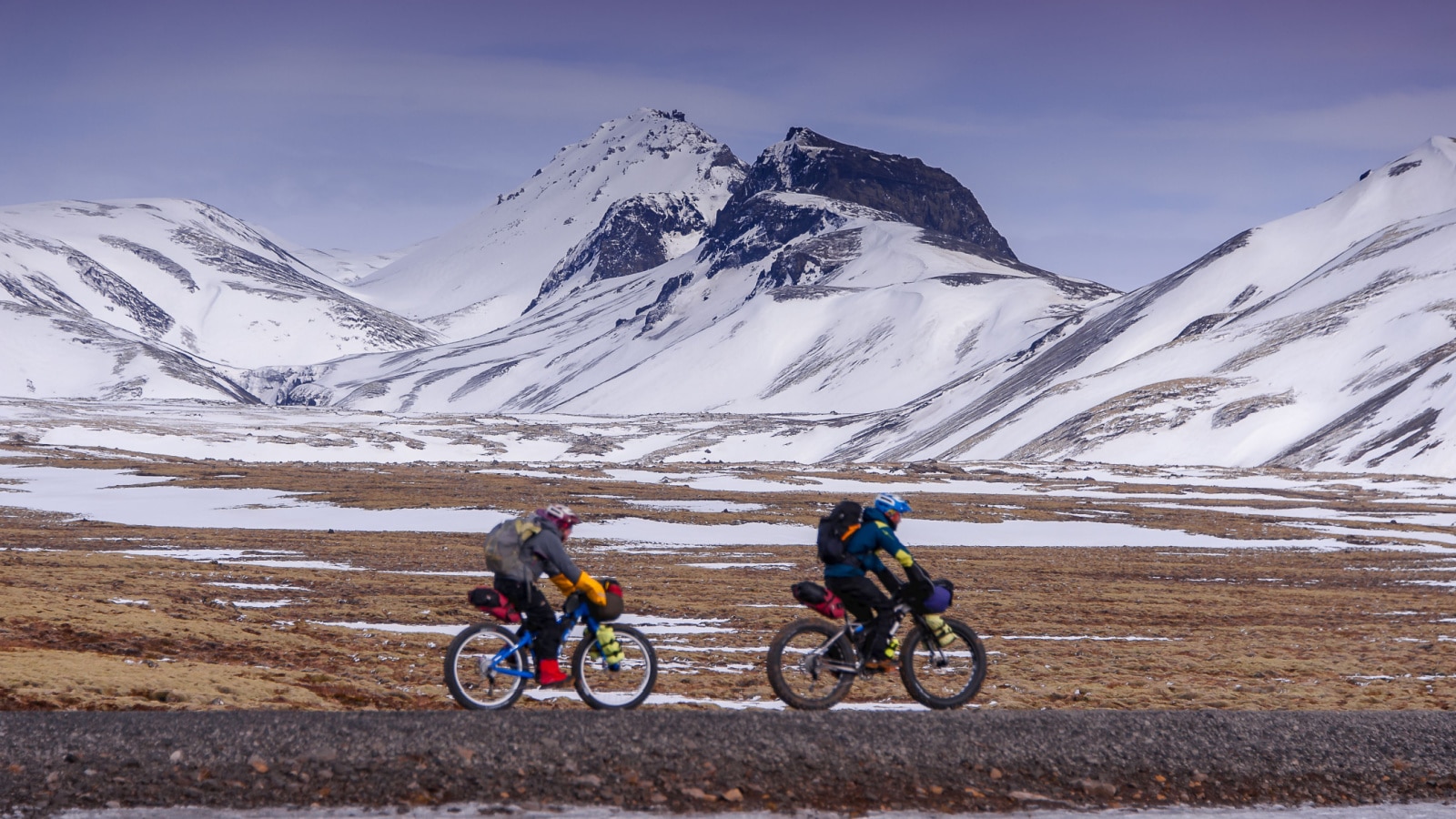 Two people cycle through a snow covered, mountainous landscape in Western Iceland