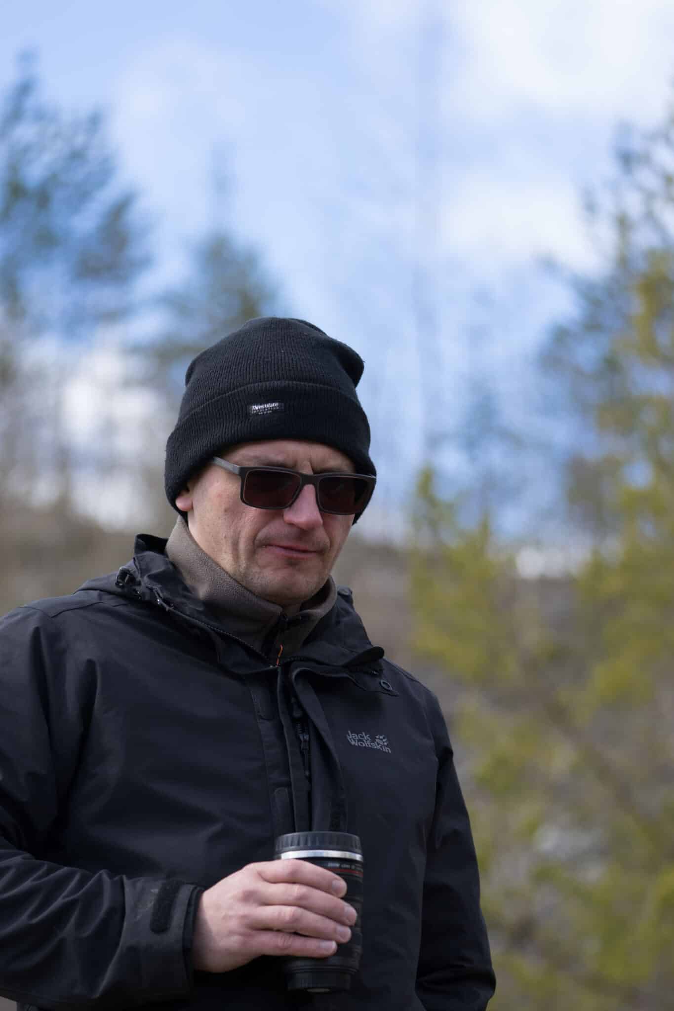 Aidu Estonia - April 30 2017: Middle aged man in black Jack Wolfskin jacket, woolen Thininsulate hat and dark sunglasses standing He has thermo mug with warm drink in his hand. Early spring background