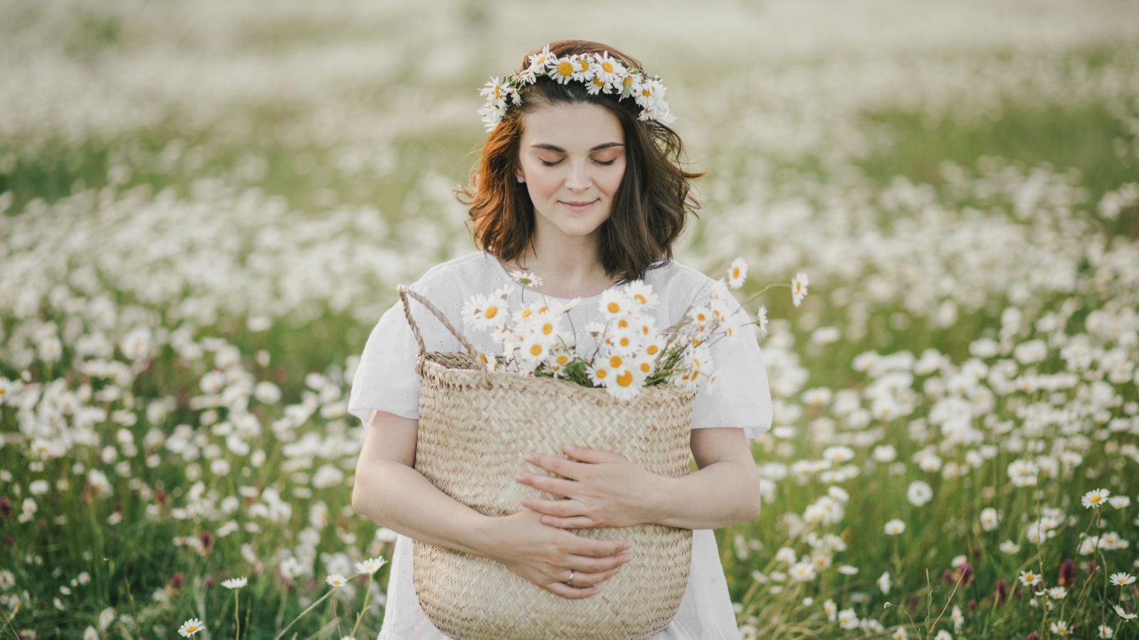 Young woman wearing white dress holding straw basket with flowers on chamomile field. Cottagecore aesthetic.