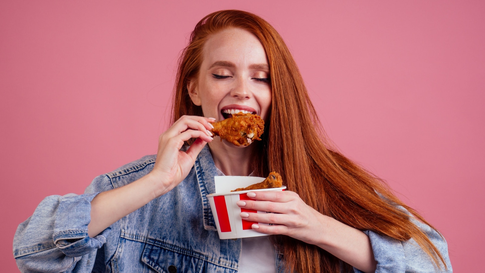 pretty long redhead ginger girl eating fries chicken in studio pink background