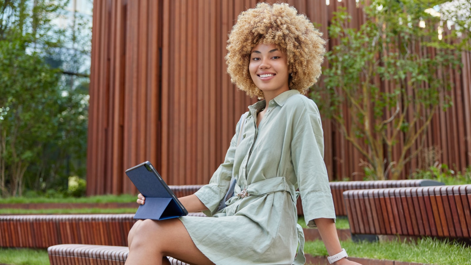 Cheerful beautiful woman wears linen dress uses modern digital tablet for reading book or browsing web pages sits outdoors on wooden bench has happy expression. Modern technologies and lifestyle