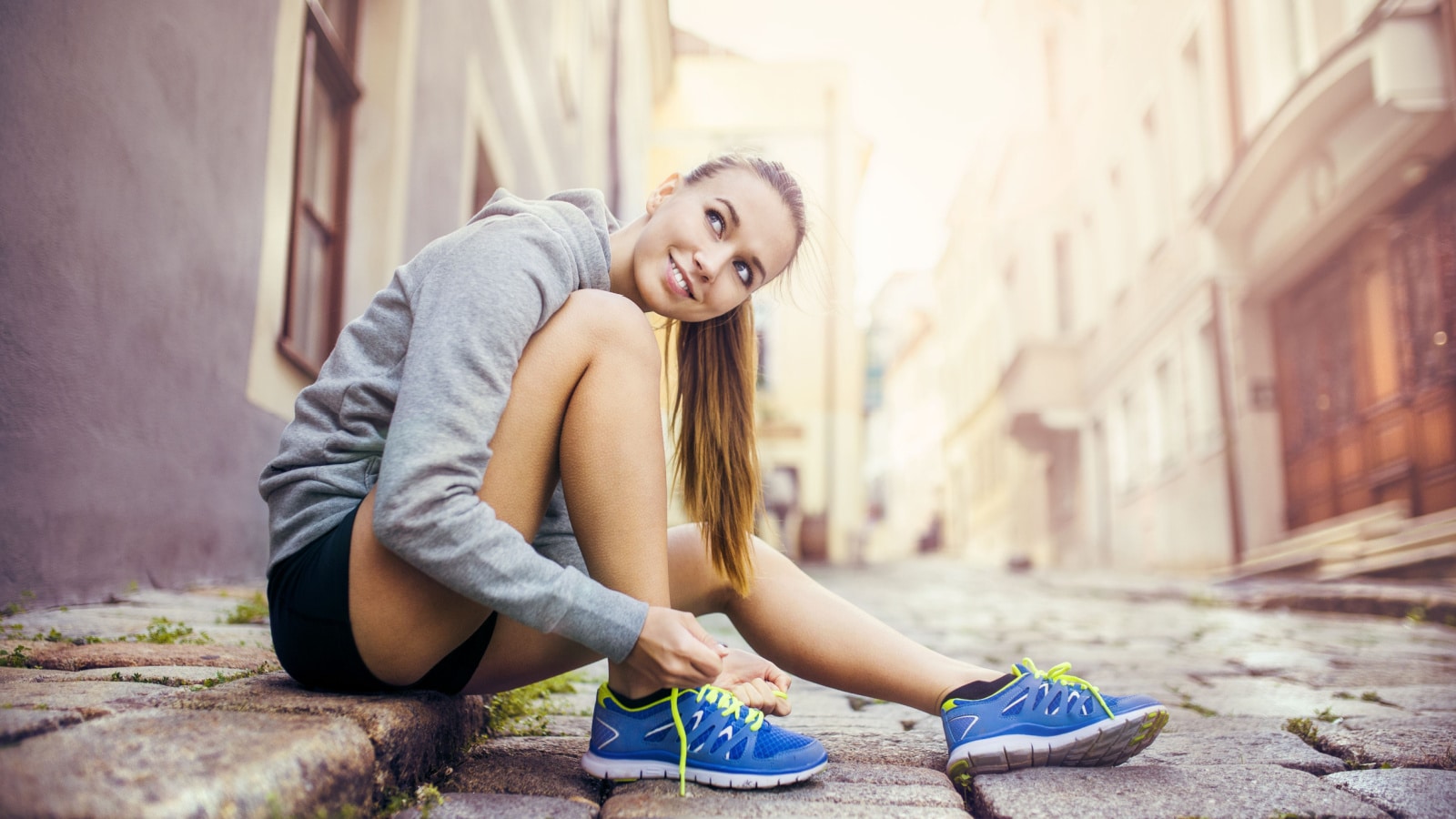 Young female runner is tying her running shoes on tiled pavement in old city center