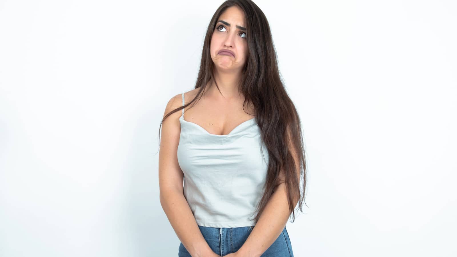 Dissatisfied young brunette woman wearing white tank top over white studio background purses lips and has unhappy expression looks away stands offended. Depressed frustrated model.