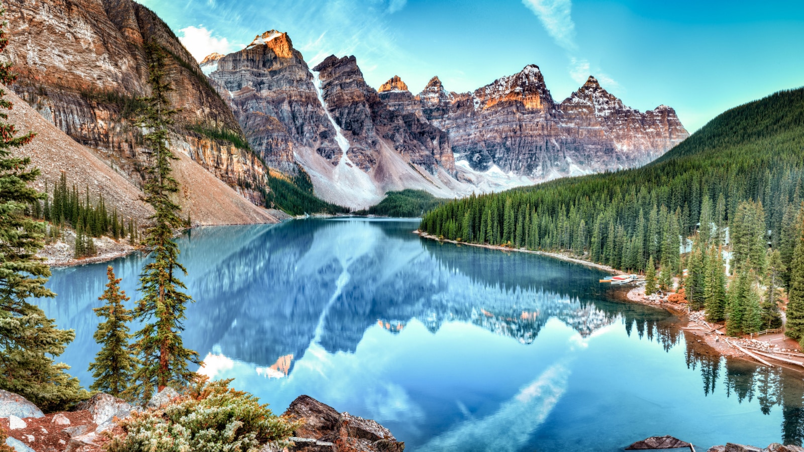 A stunning panorama image of Moraine lake panorama in Banff National Park, Alberta, Canada. Mountain ridges loom in the background, over a winding lake and hillsides covered in trees. The mountains reflect in the crystal clear waters.