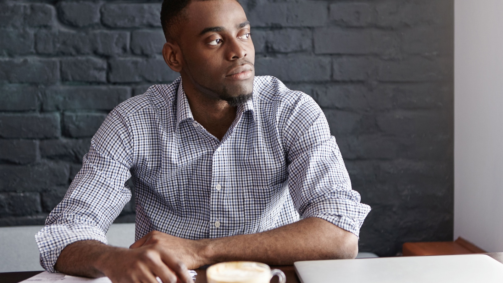 African-Ameican entrepreneur wearing shirt with rolled up sleeves looking through window with thoughtful and serious face expression, feeling nervous before meeting with business partners at cafe
