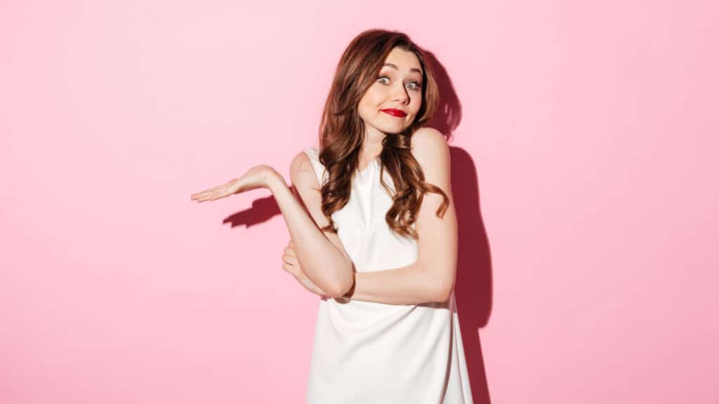 Portrait of a uncertain pretty woman shrugging shoulders isolated over pink background