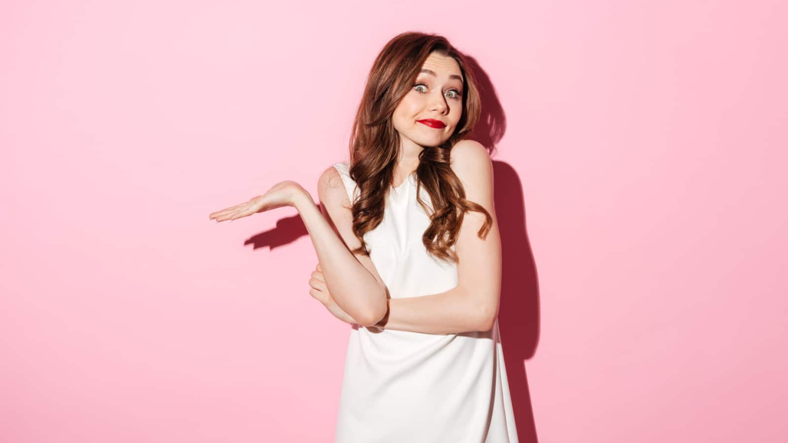 Portrait of a uncertain pretty woman shrugging shoulders isolated over pink background