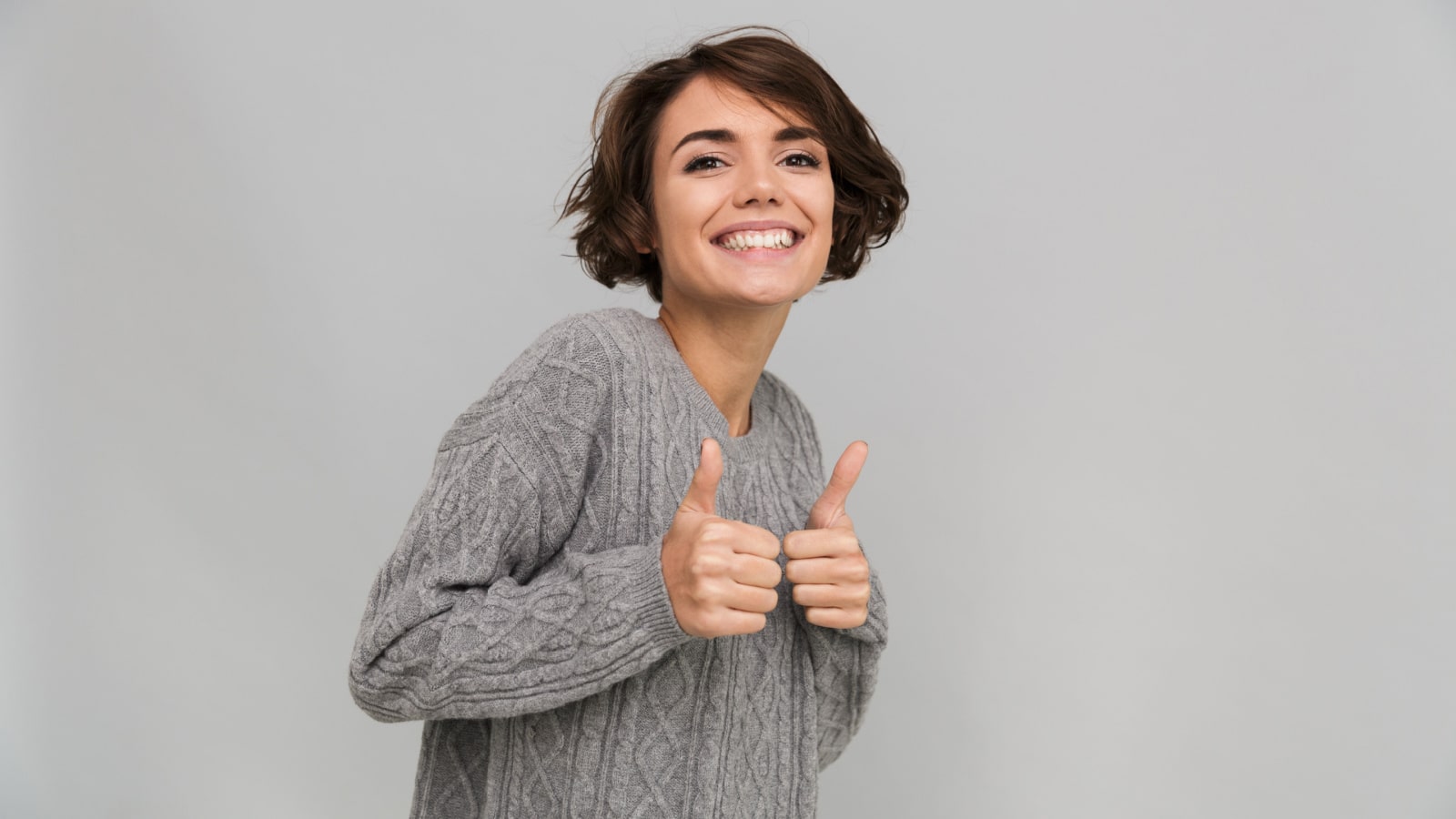 Image of happy young lady dressed in sweater standing isolated over grey background. Looking camera showing thumbs up gesture.