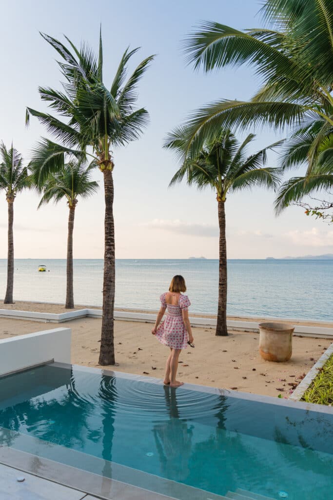Young woman in a dress standing on the edge of a pool looking out at the beach surrounded by palm trees in Thailand