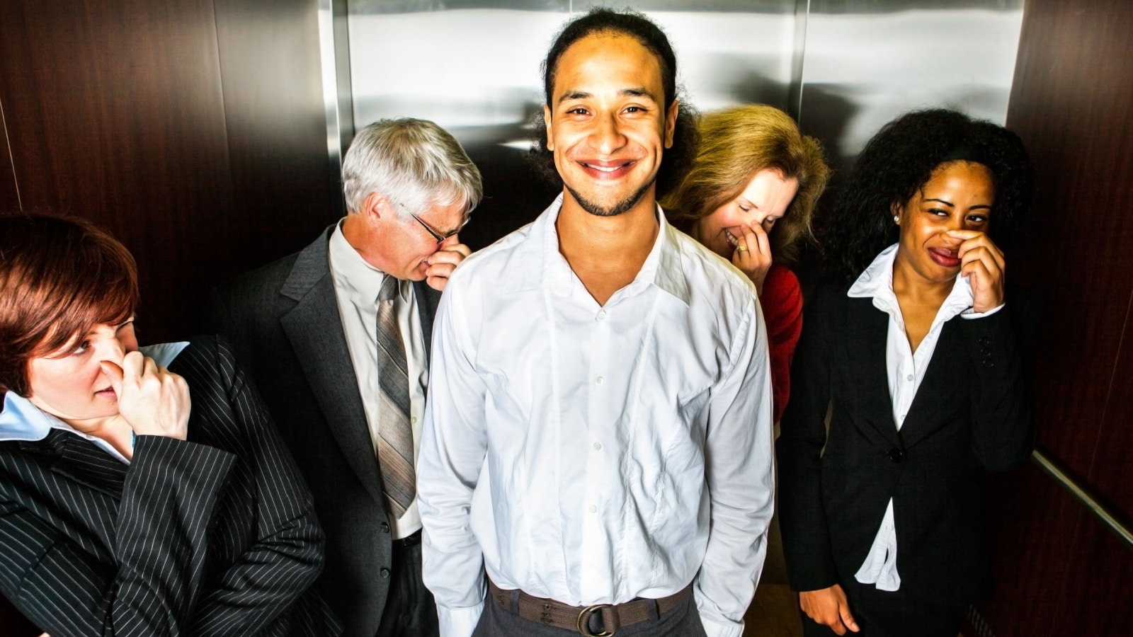 A young man smiles in embarrassment in an elevator while people hold their noses.
