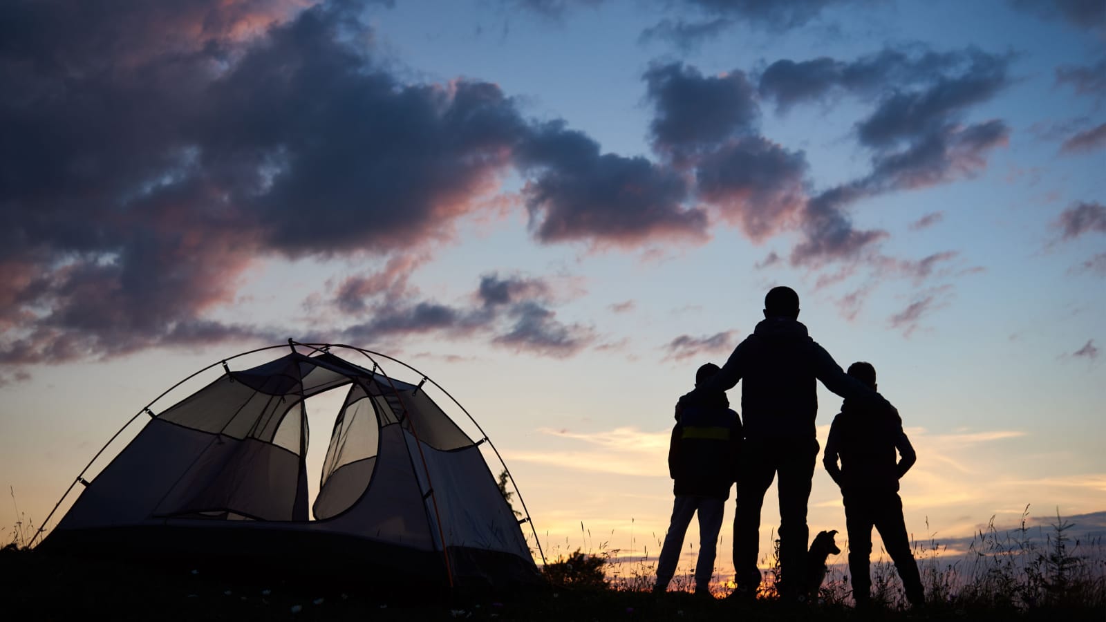 Rear view of silhouette of a father backpacker hugging two children. Tourists guys and dog stands beside camping on the top of mountain near tent at sunset under evening sky with clouds.