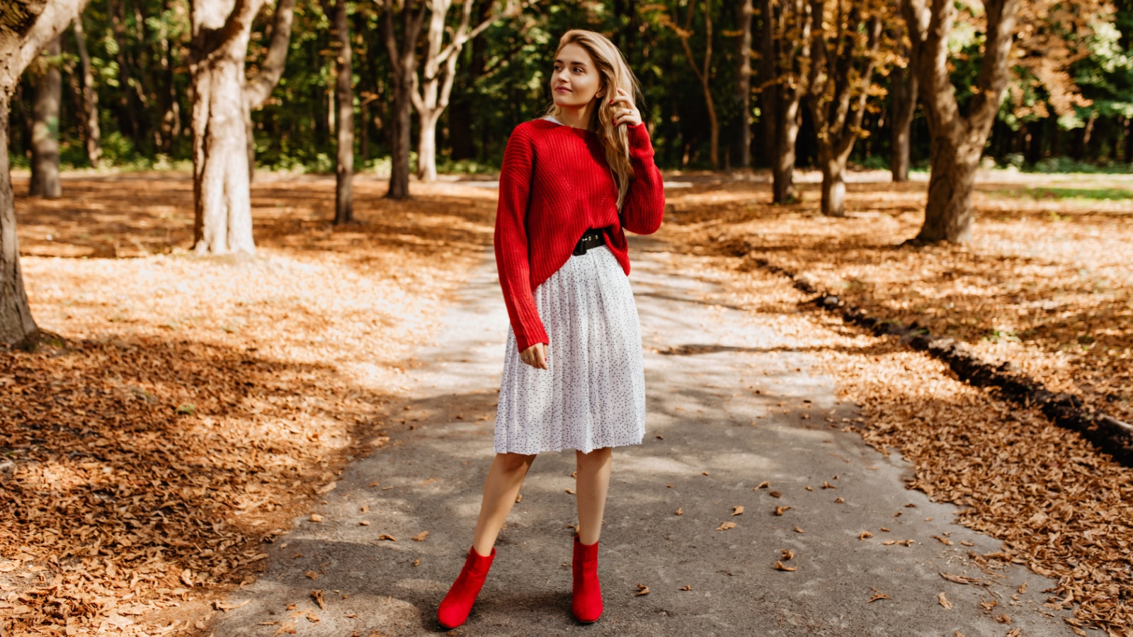 Attractive blonde girl posing in trendy clothes in the autumn park. Young woman wearing red sweater and white skirt outdoor.