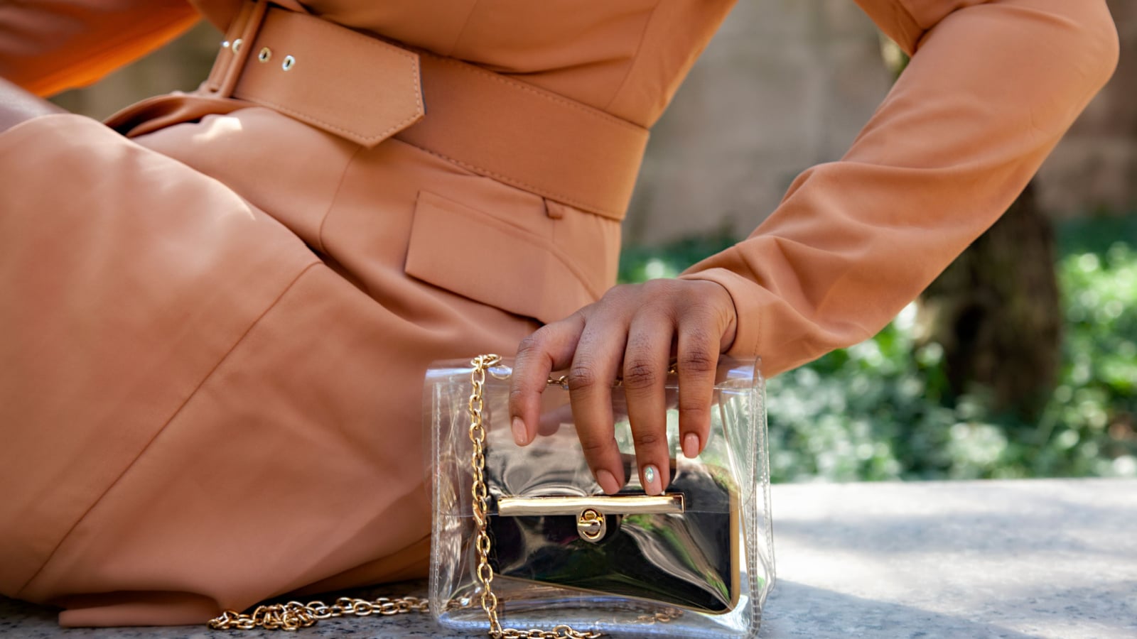 close up shop of a African american black woman hands holding a clear hand bag by a water fountain in Chicago. she's wearing a fashionable brown trench coat.