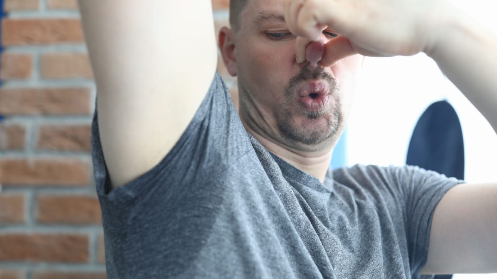 Portrait of middle-aged man holding arm up and closing nose with hands because of bad smell. Sweaty grey shirt. Male after run or sport workout. Personal hygiene concept