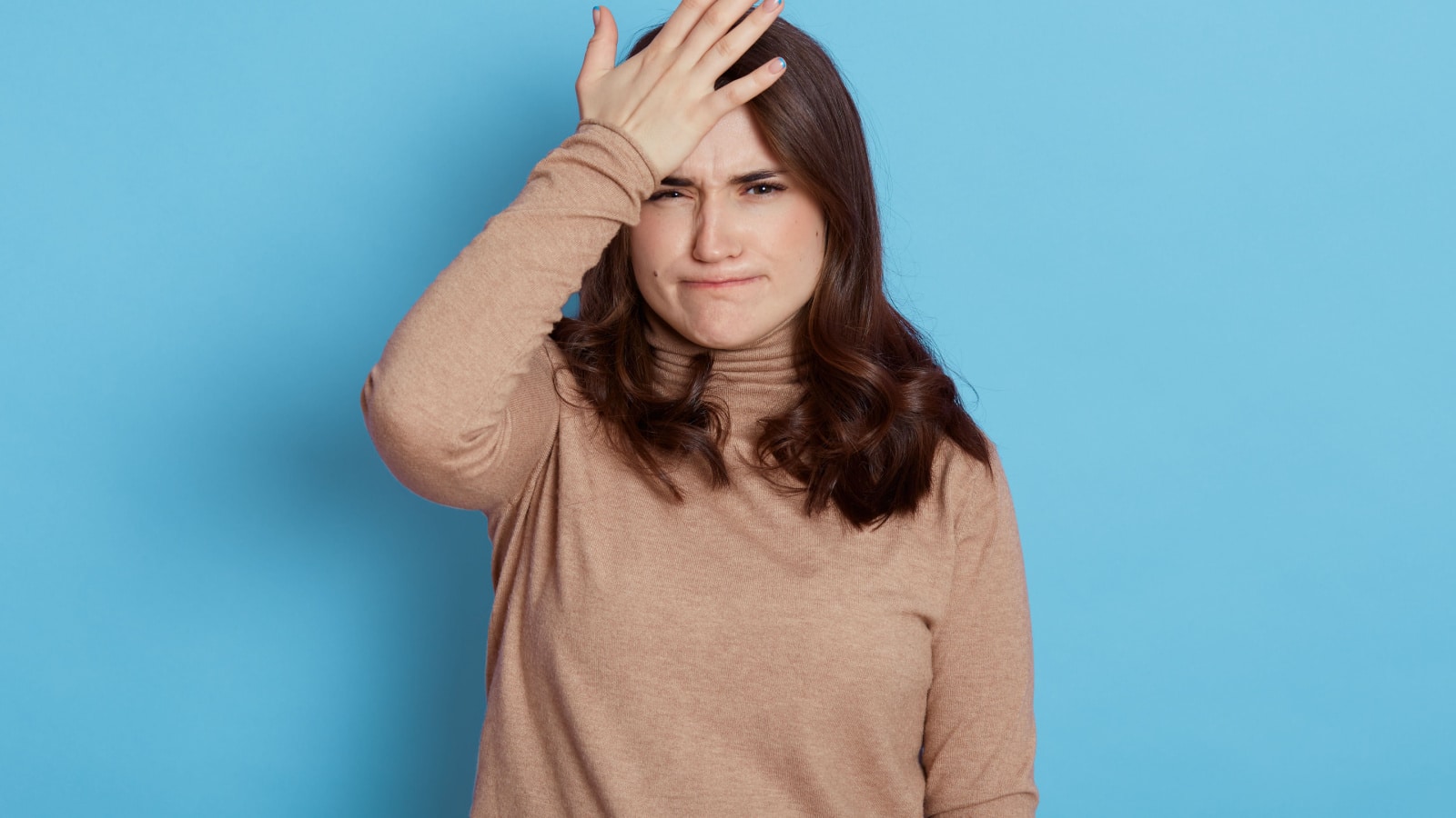 Disappointed woman keeps palm on forehead, regrets, did something wrong, having problematic situation, dressed in casual outfit, poses on blue wall, forgets about important task, lady with pursed lips