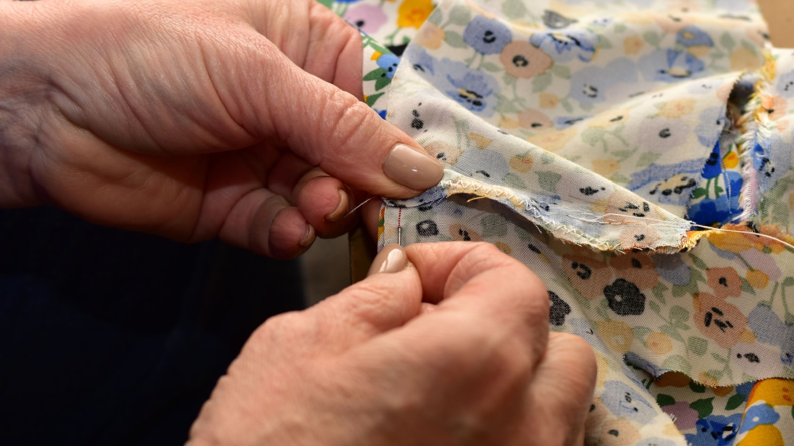 The picture shows the hands of a woman who are sewing two pieces of fabric with a needle and thread.