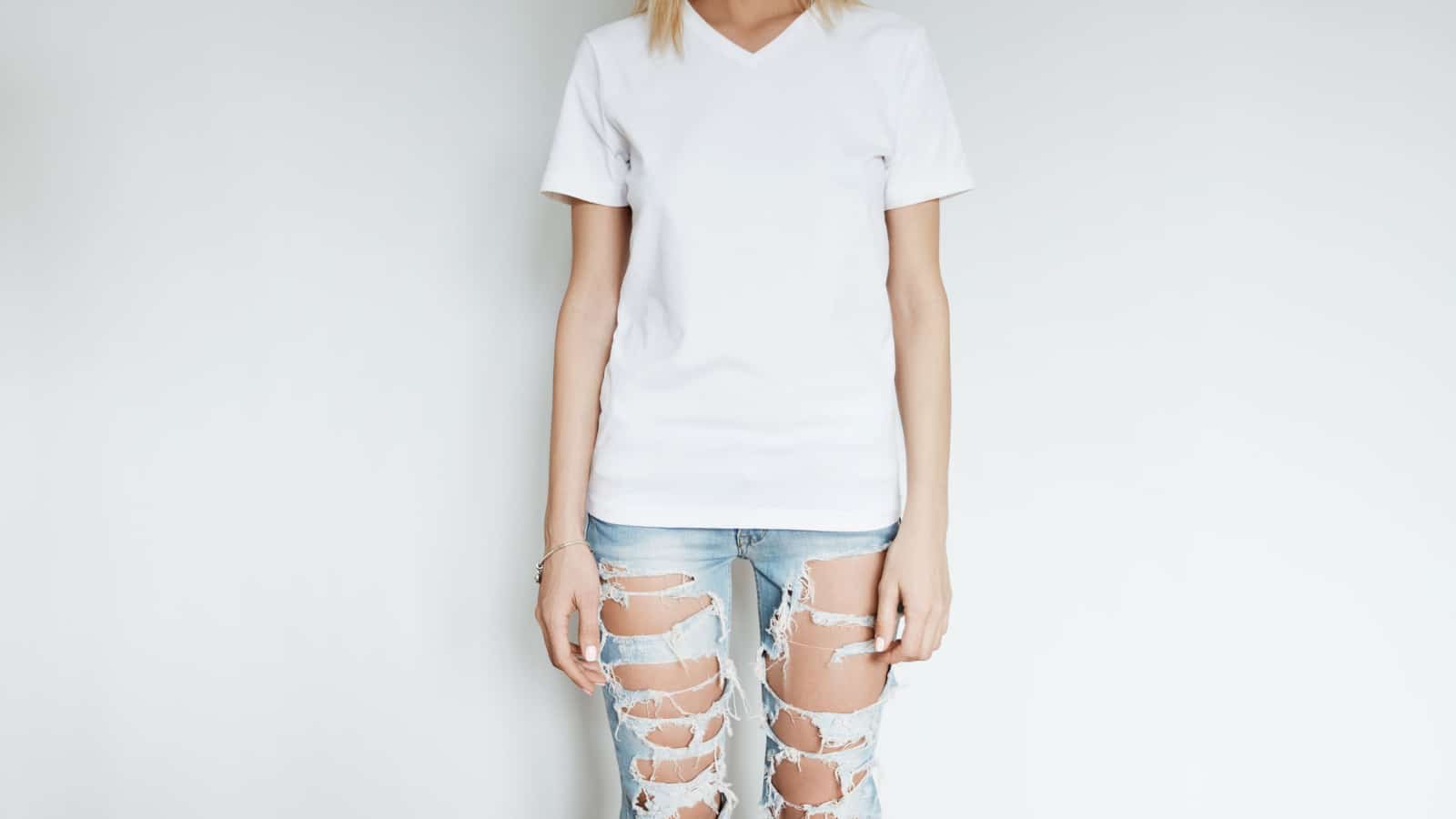 Cropped view of blonde slim girl wearing blank T-shirt with copy space for your content. Young woman in ripped jeans posing isolated against blank white wall. Advertising and T-shirt design concept