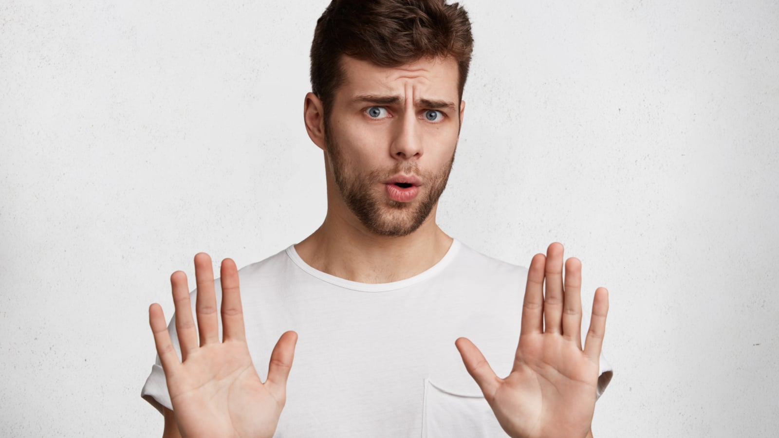 Attractive brunet male shows refusal gesture, doesn`t want to participate in meeting, says: It`s not for me, leave me in piece, has angry expression, poses against white concrete studio background
