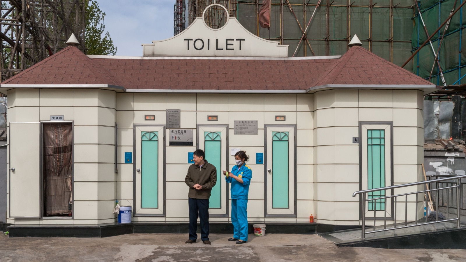 Beijing, China - April 26, 2010: Neat beige with brown roof modern public toilet cabin along street. Maintenance worker in blue uniform on site. Man in photo, too.