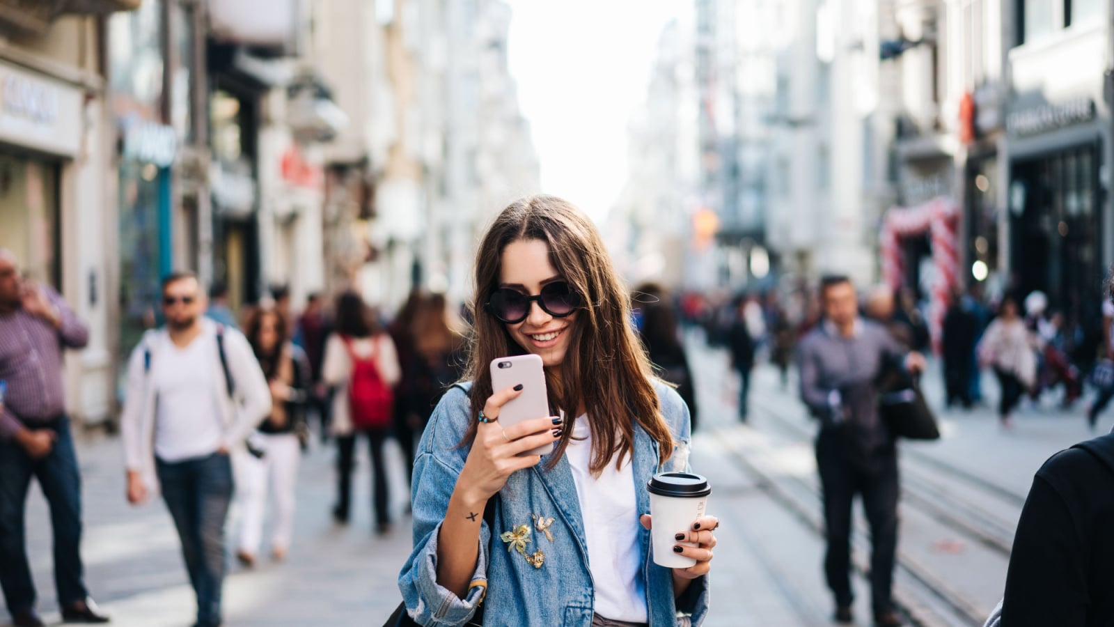 Beautiful smiling woman walking on crowded city street from work with coffee cup and texting on mobile phone