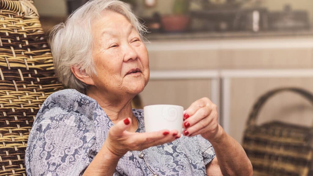 Old lady (japanese descendant) seated comfortably at home holding a cup of tea with a happy expression of who is having a good conversation or telling stories.