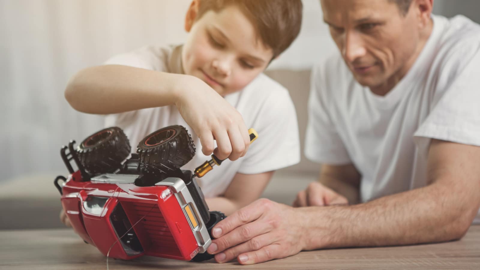 Interested boy is repairing toy by instrument with help of his dad. Focus on small red car