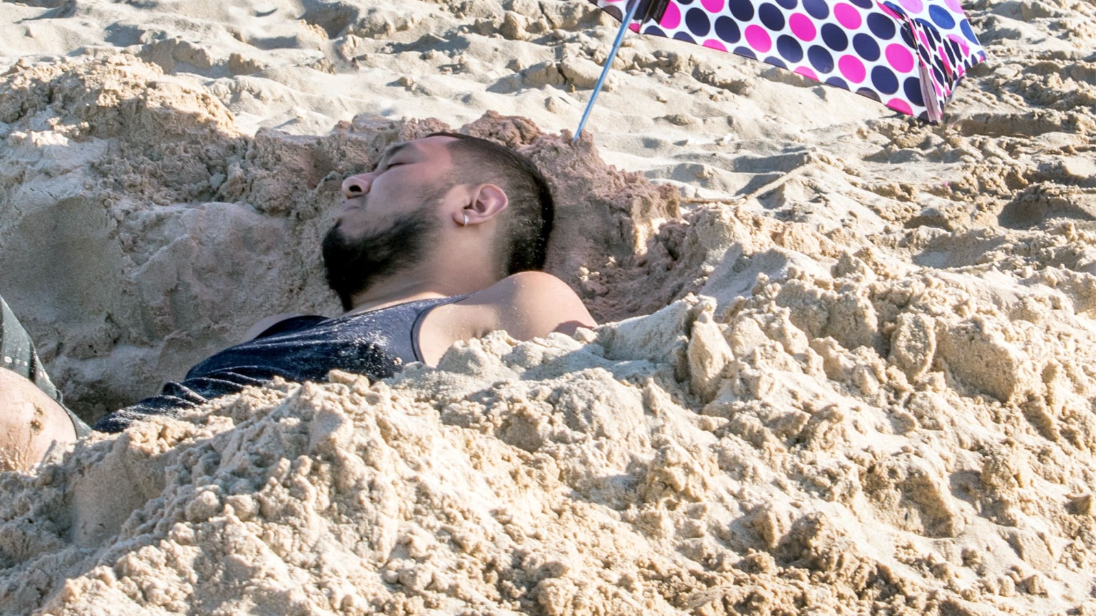 St Joseph MI USA , May 27 2018; A tired man relaxes in a cool hole dug in the sand, and under a shady polka dot umbrella