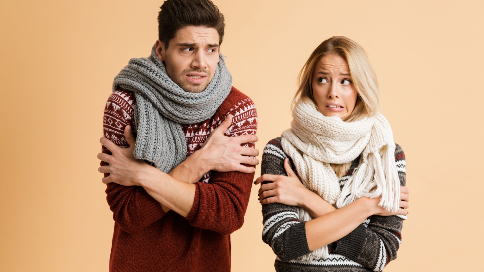 Portrait of a frozen young couple dressed in sweaters and scarves standing together isolated over beige background, shaking, looking at each other