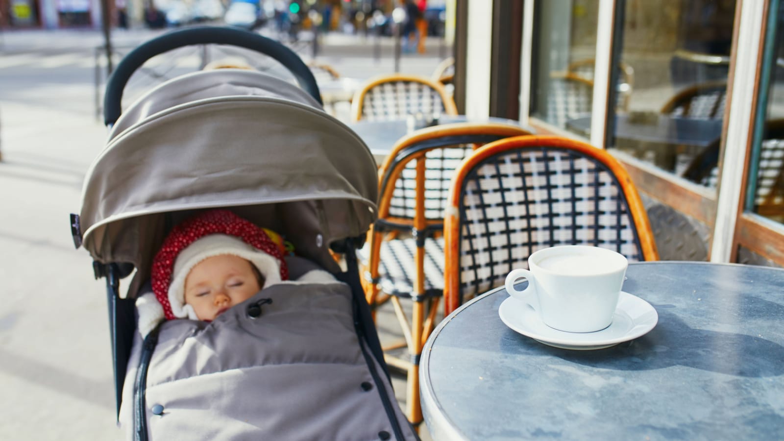 Baby girl sleeping in pram on outdoor terrace of Parisian street cafe with cup of hot coffee on the table. Going out with kids