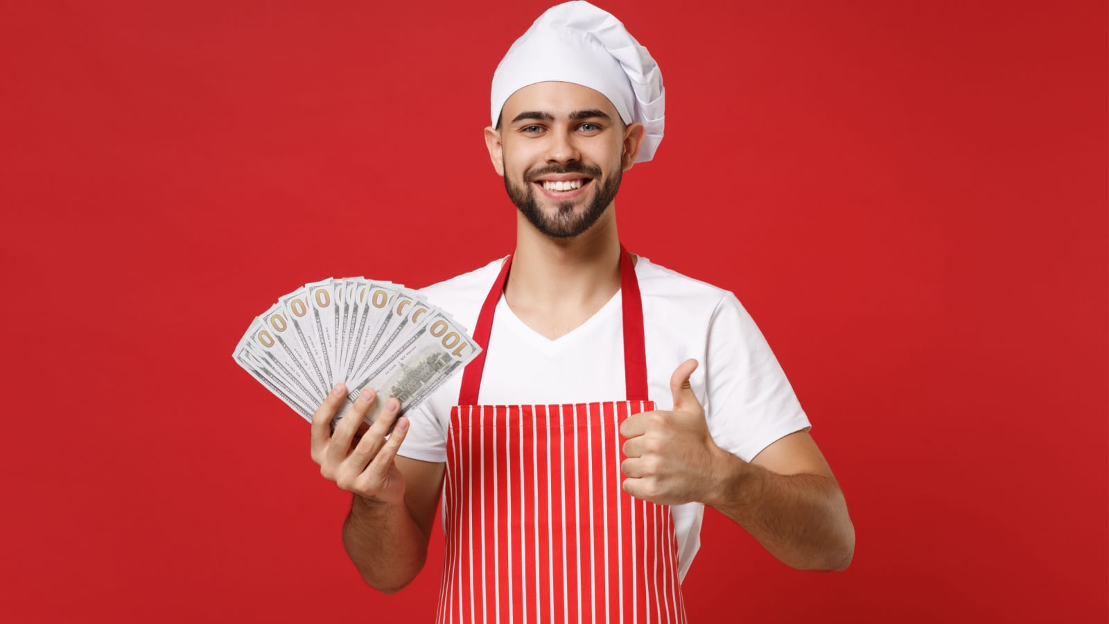 Smiling young bearded male chef cook or baker man in striped apron toque chefs hat isolated on red background. Cooking food concept. Mock up copy space. Holding fan of cash money, showing thumb up
