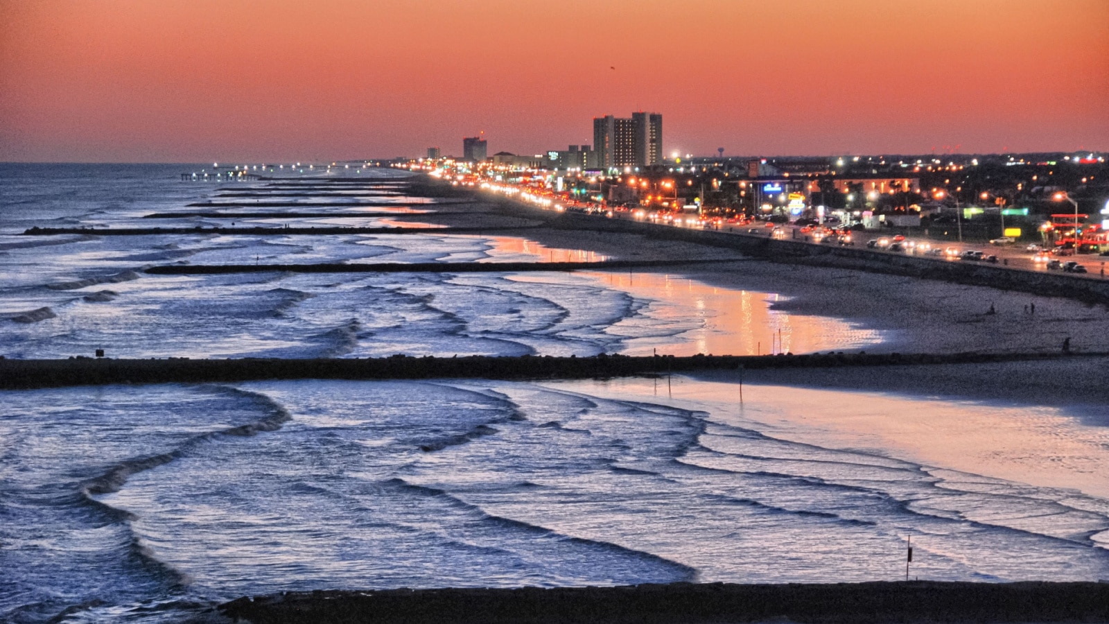 An aerial photo of Galveston Texas along the shoreline, with waves breaking along the beach at dusk, and the city lights in the background.