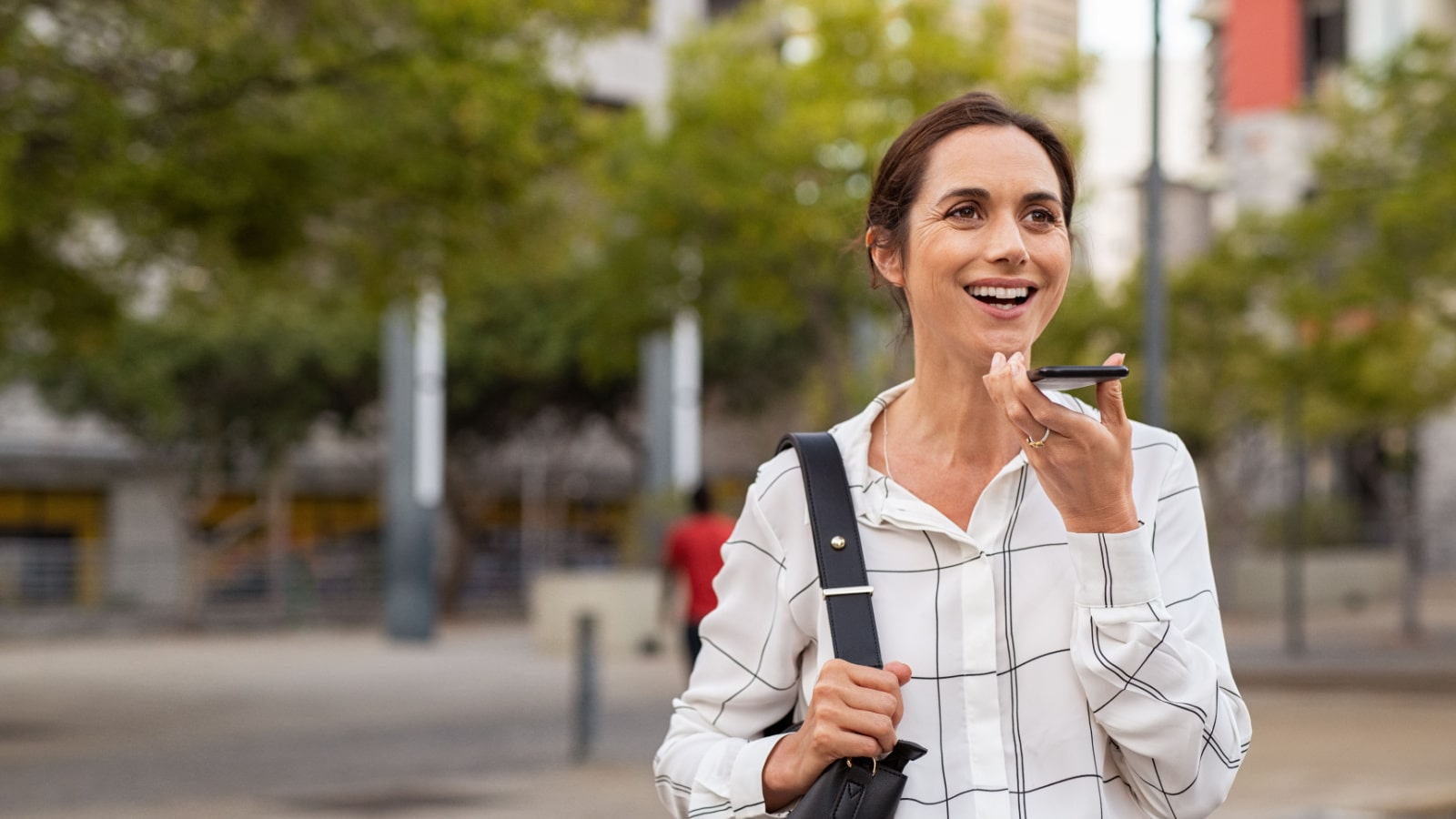 Smiling mature woman using vocal assistance over smartphone. Successful entrepreneur using mobile phone over loudspeaker on street. Happy businesswoman using cellphone for vocal message, copy space.