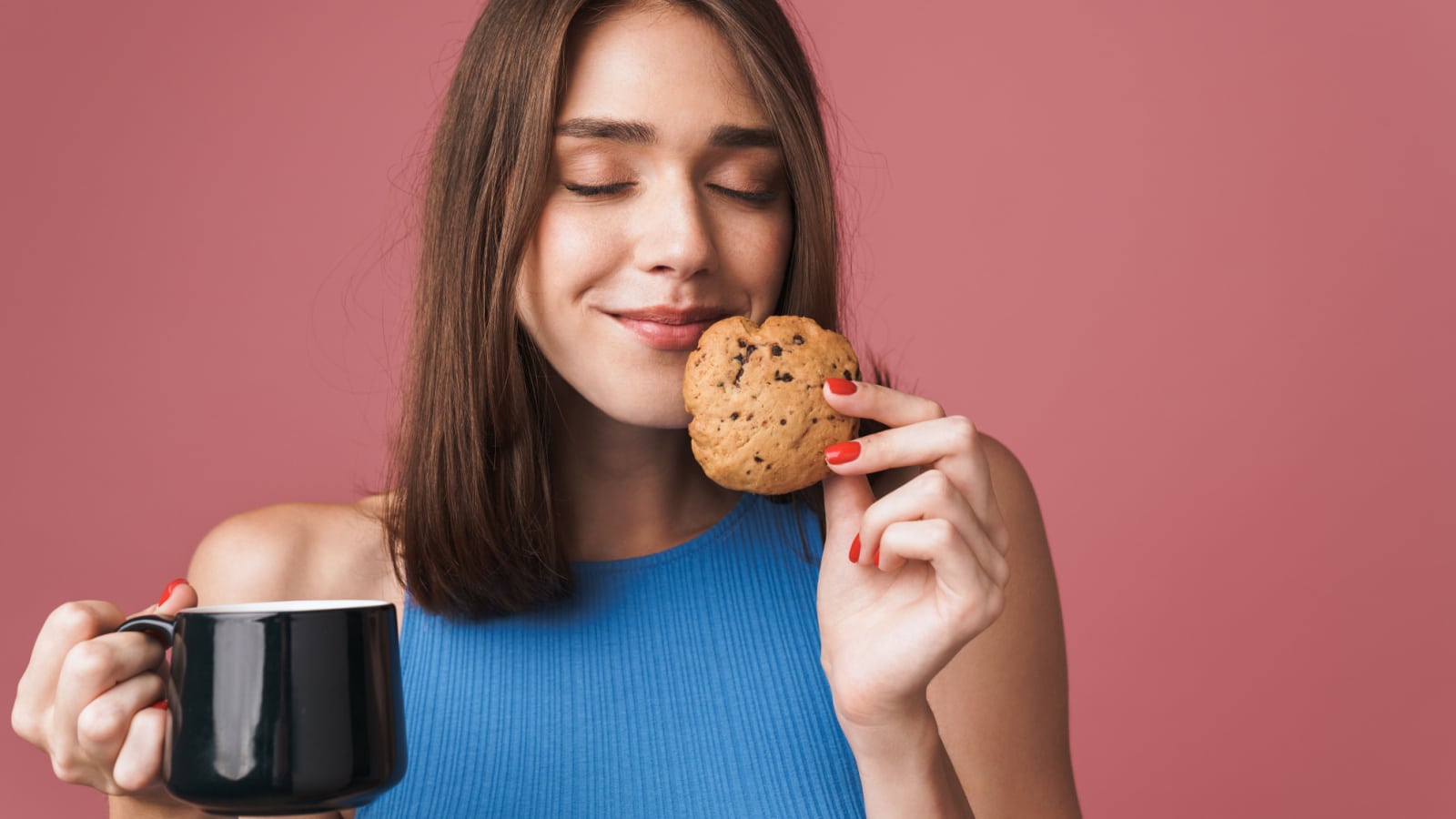 Portrait of a young smiling attractive brunette woman standing isolated over pink background, holding a cup of coffee and eating a chocolate chip cookie