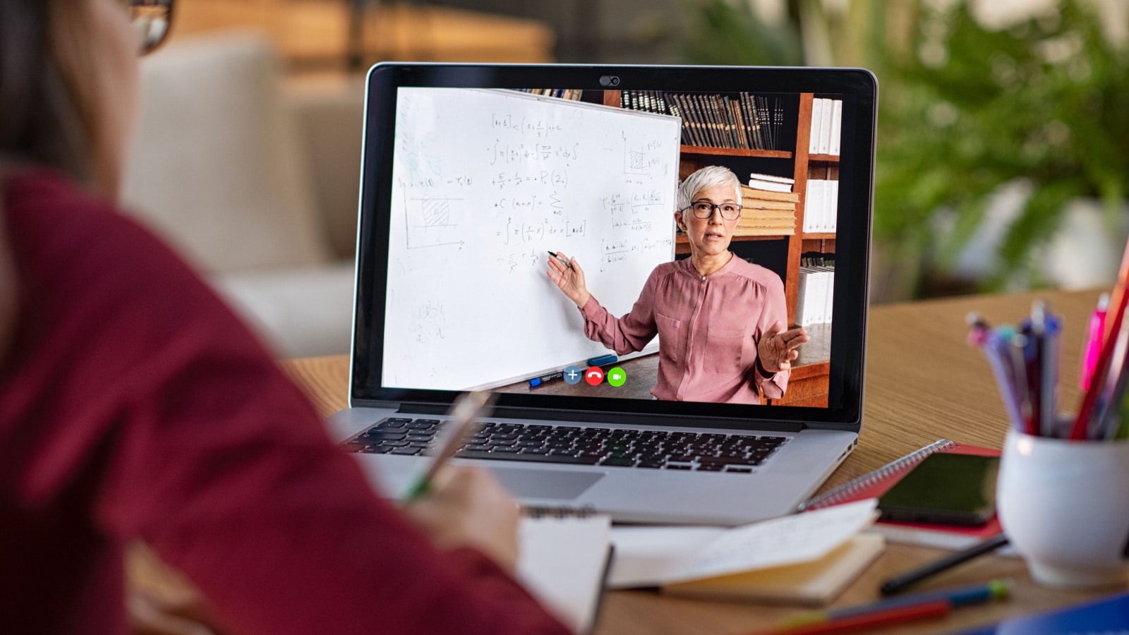 Young student watching lesson online and studying from home. Young woman taking notes while looking at computer screen following professor doing math on video call. Girl studying from home on pc.