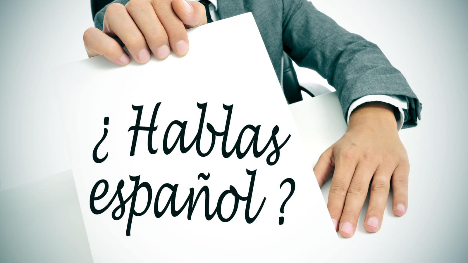 a man wearing a suit holding a signboard with the sentence hablas espanol? do you speak spanish? written in spanish on it