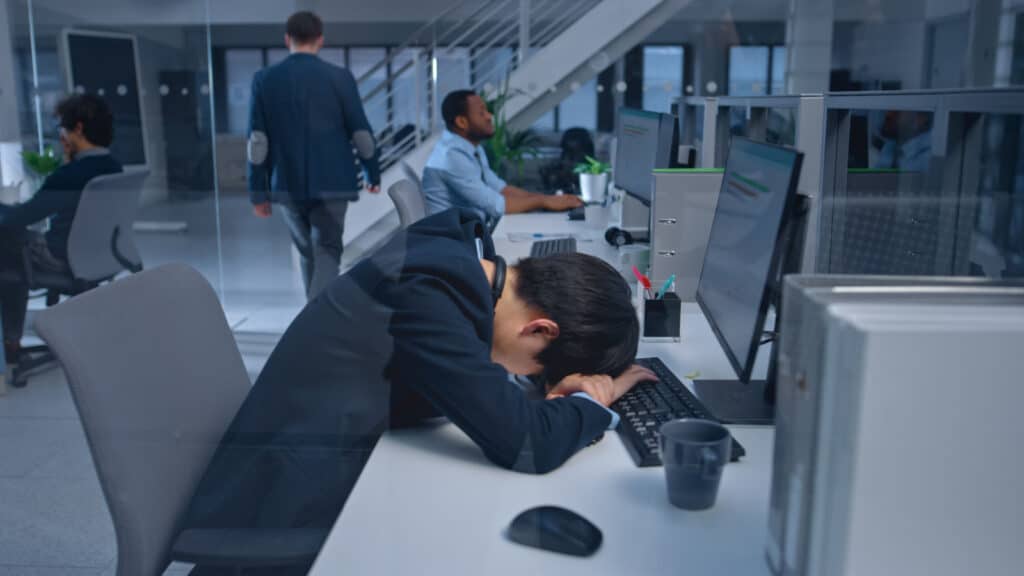 Young Japanese Manager is Tired at his Work Place with Desktop Computer and Falls Asleep. Diverse Multi-ethnic Business People Work on Computers in Modern Open Office.