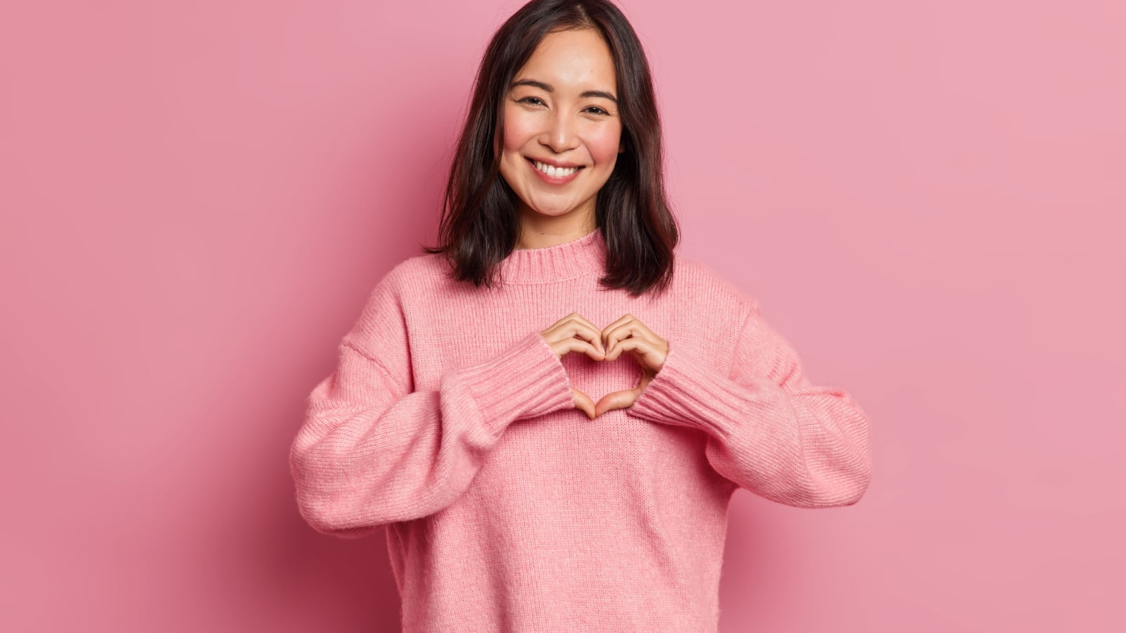 Attractive brunette young Asian woman feels happy and romantic shapes heart gesture expresses tender feelings wears casual jumper poses against pink background. People affection and care concept