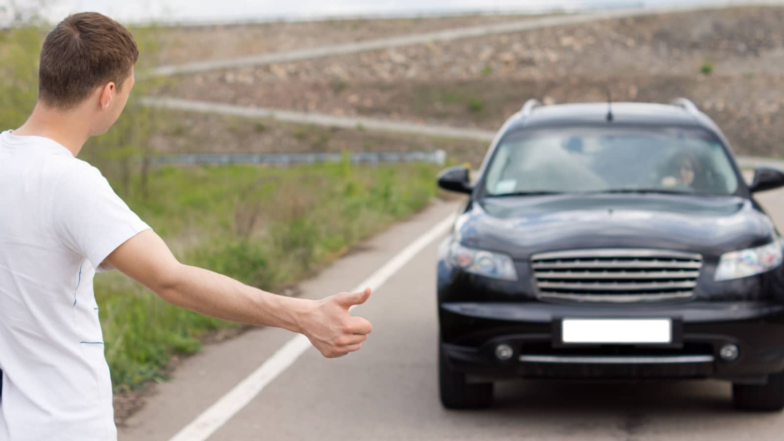 Young man hitching a lift on a country road standing at the side of the road with his thumb out flagging down a passing car