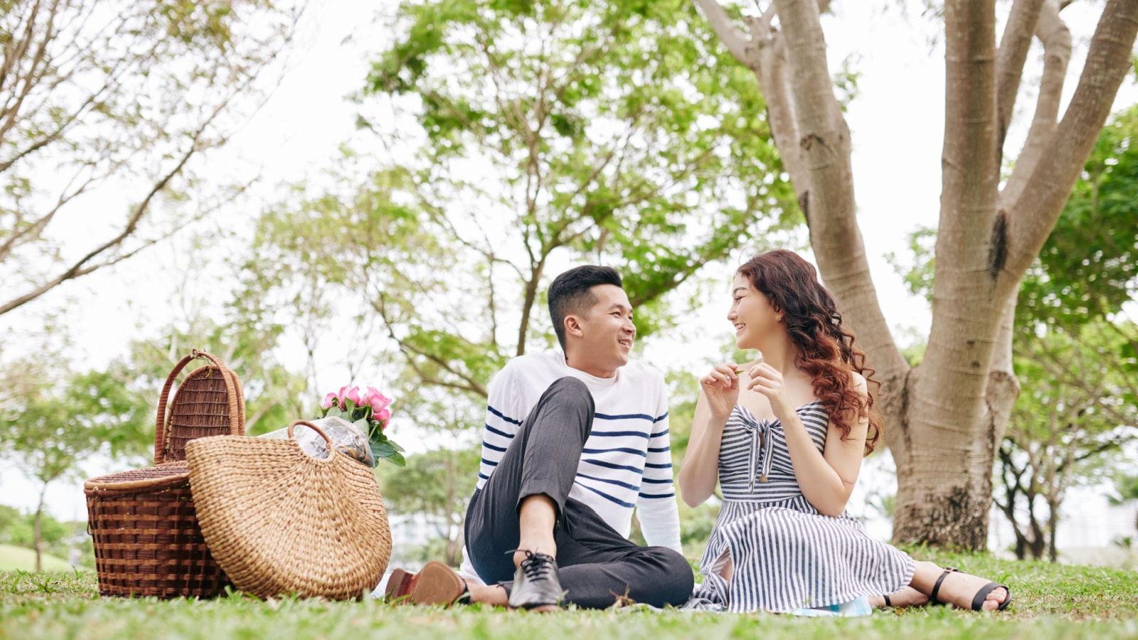 Cheerful young Asian couple in love resting on blanket in park and talking