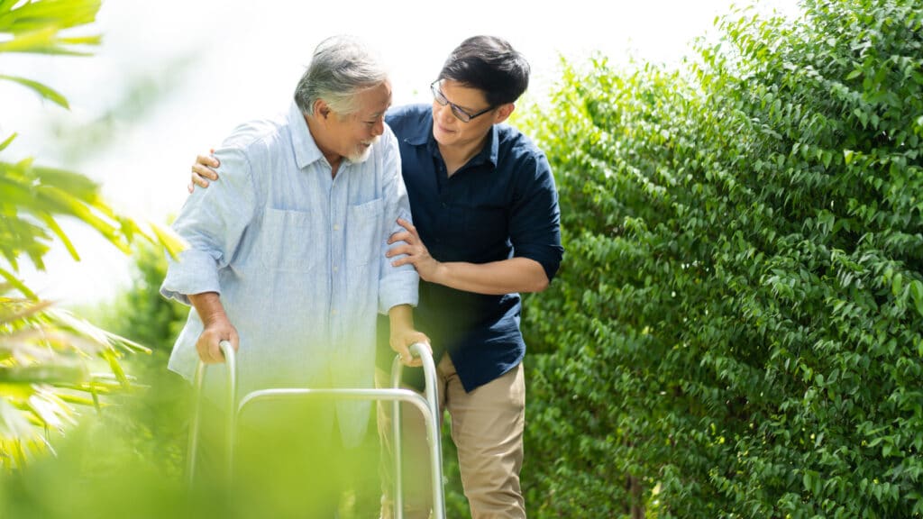 Elderly Asian father and Adult son walking in backyard. Positive Asian man caregiver helping patient