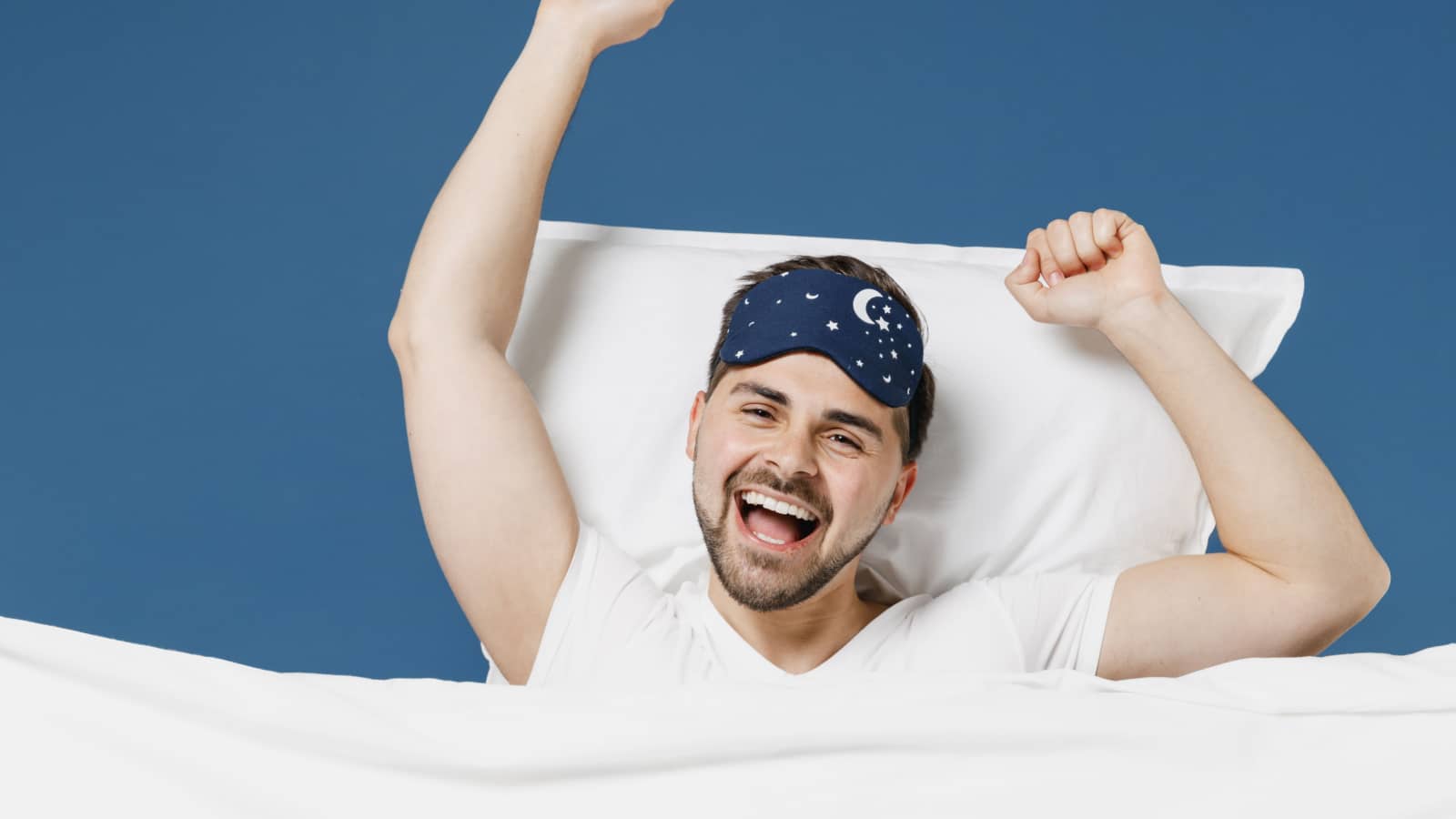 Young happy man 20s wear pajamas jam sleep mask rest relaxing home with lies wrap covered under blanket duvet with look camera yawning isolated on dark blue background Good mood night bedtime