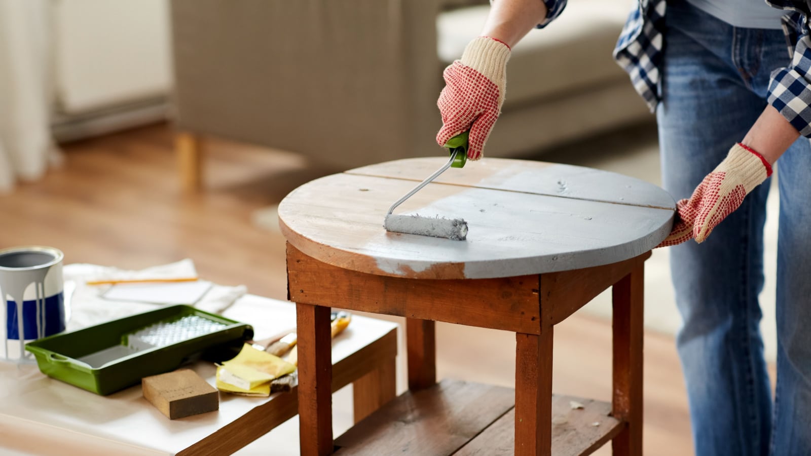 furniture renovation, diy and home improvement concept - close up of woman in gloves with paint roller painting old wooden table in grey color