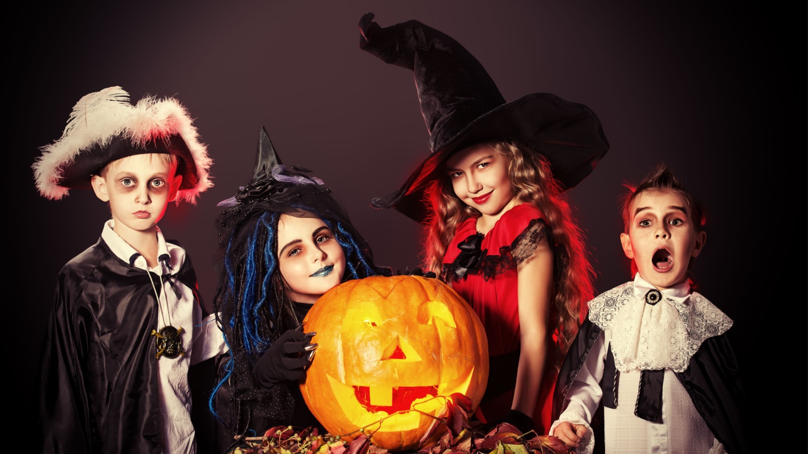 Cheerful children in halloween costumes posing with pumpkin over dark background.