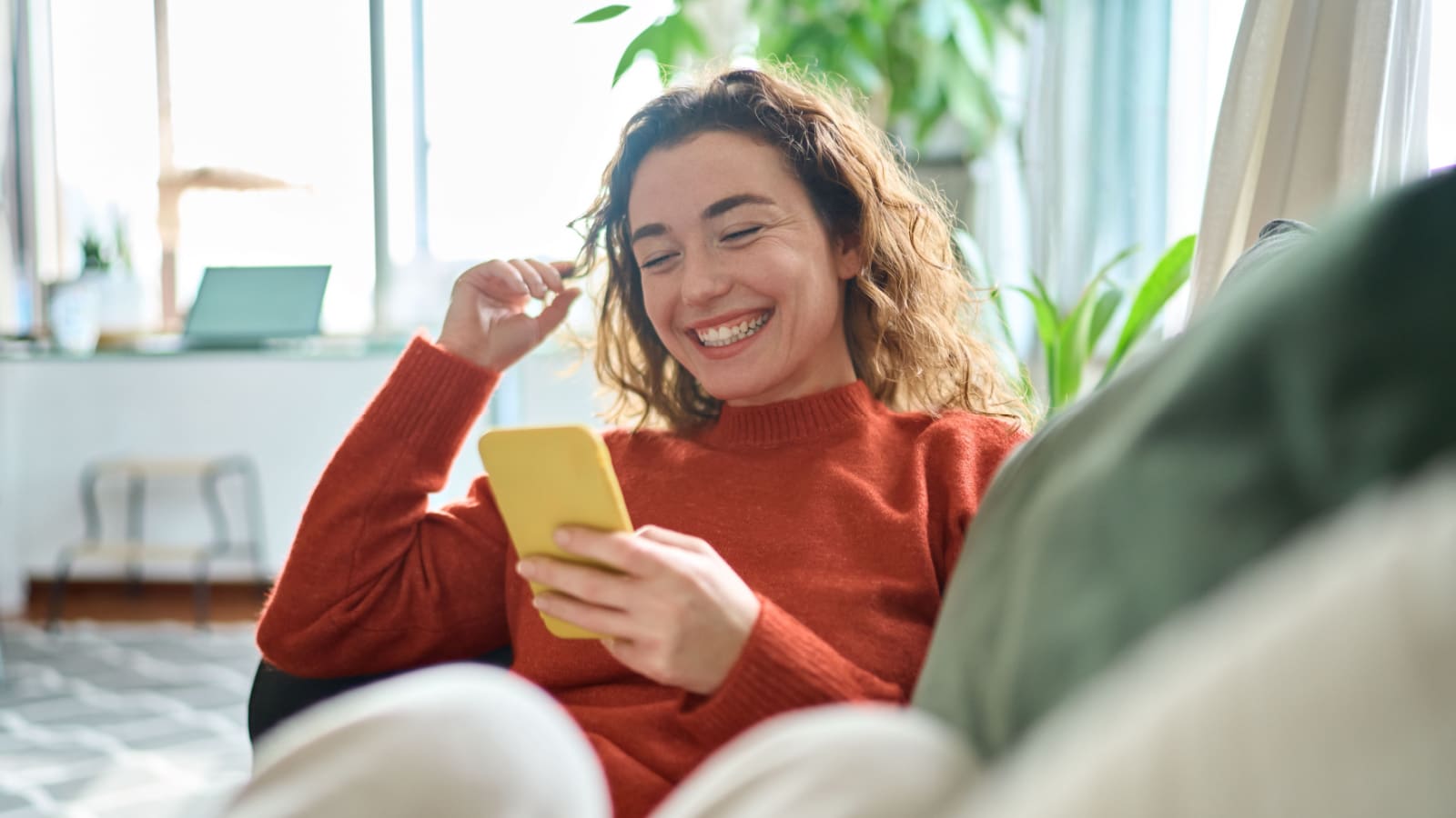 Happy relaxed young woman sitting on couch using cell phone, smiling lady laughing holding smartphone, looking at cellphone enjoying doing online ecommerce shopping in mobile apps or watching